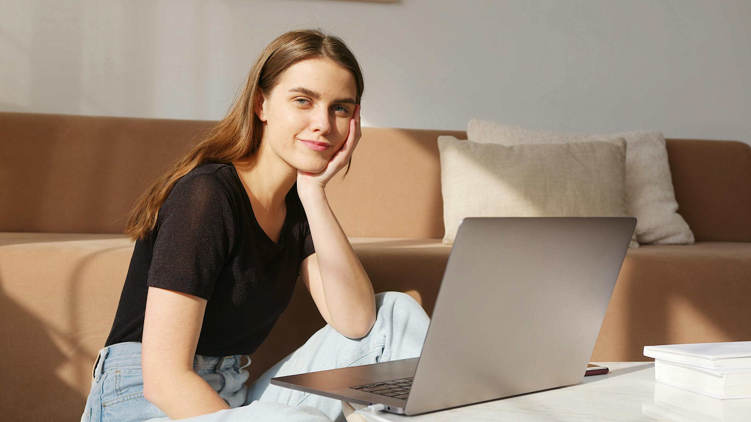 FAQs Positive glad young lady in casual clothes sitting on floor and browsing modern netbook while leaning head on hand and looking at camera happily
